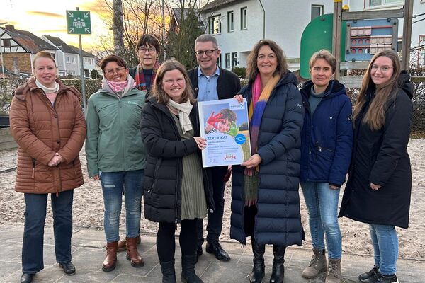 Eine Gruppe von Kita-Mitarbeitenden steht draußen, im Hintergrund sind Häuser zu sehen. In der Mitte stehen zwei Frauen, die ein DIN A3 großes Plakat in den Händen halten.