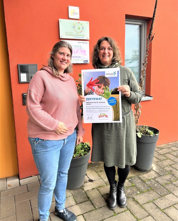 Zwei Frauen vor einer orangenen Wand. Die rechte Frau übergibt der linken ein Zertifikat.