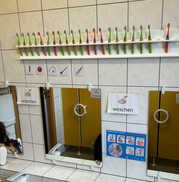 Das Foto zeigt eine Wand eines Waschraums in einer Kita mit diversen Plakaten zum richtigen Händewaschen. Oben gibt es eine Leiste mit einzeln aufgehängten Kinderzahnbürsten.